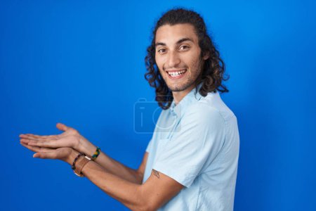 Photo for Young hispanic man standing over blue background pointing aside with hands open palms showing copy space, presenting advertisement smiling excited happy - Royalty Free Image