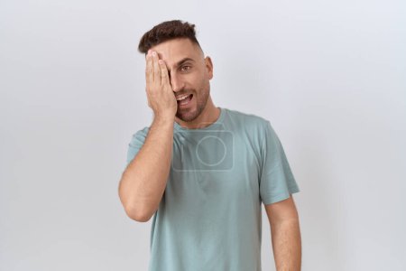 Photo for Hispanic man with beard standing over white background covering one eye with hand, confident smile on face and surprise emotion. - Royalty Free Image
