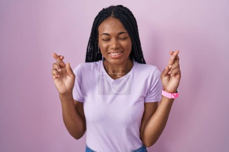 Téléchargez les photos : Femme afro-américaine avec des tresses debout sur fond rose geste doigt croisé souriant avec espoir et les yeux fermés. chance et concept superstitieux. - en image libre de droit
