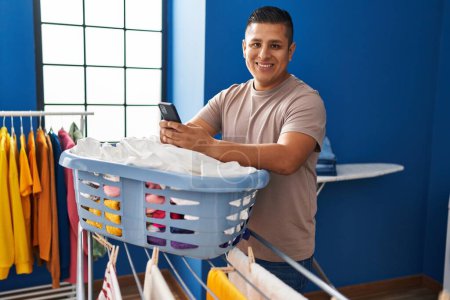 Foto de Joven latino usando smartphone colgando ropa en tendedero en la lavandería - Imagen libre de derechos