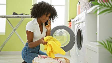 Téléchargez les photos : Femme afro-américaine parlant sur smartphone laver les vêtements souriant à la buanderie - en image libre de droit
