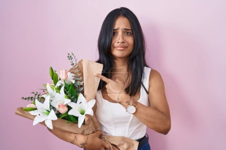 Foto de Mujer morena sosteniendo ramo de flores blancas señalando a un lado preocupado y nervioso con el dedo índice, preocupado y expresión sorprendida - Imagen libre de derechos