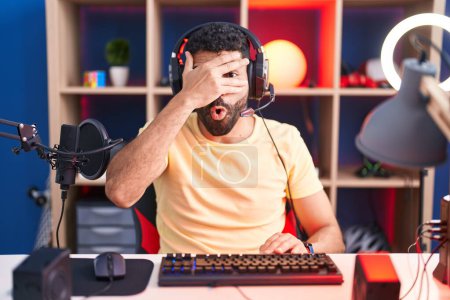 Téléchargez les photos : Homme hispanique avec barbe jouant à des jeux vidéo avec écouteurs regardant dans le choc couvrant le visage et les yeux avec la main, regardant à travers les doigts avec une expression embarrassée. - en image libre de droit