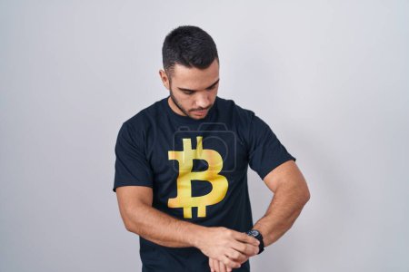 Foto de Joven hombre hispano con camiseta bitcoin comprobando la hora en el reloj de pulsera, relajado y confiado - Imagen libre de derechos