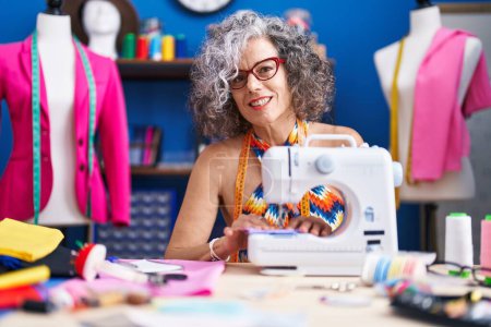 Foto de Mujer de pelo gris de mediana edad sastre sonriendo confiado utilizando la máquina de coser en el estudio de costura - Imagen libre de derechos