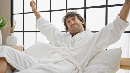 Photo for Young hispanic man waking up stretching arms wearing bathrobe at bedroom - Royalty Free Image