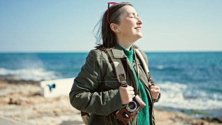 Téléchargez les photos : Femme hispanique mature avec des cheveux gris touriste portant un sac à dos souriant regardant sur le côté au bord de la mer - en image libre de droit