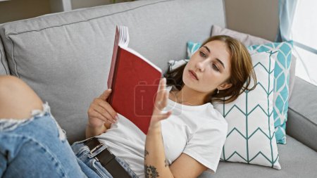 Photo for Young woman reading book lying on sofa at home - Royalty Free Image