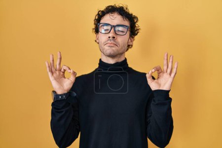 Téléchargez les photos : Hispanic man standing over yellow background relaxed and smiling with eyes closed doing meditation gesture with fingers. yoga concept. - en image libre de droit