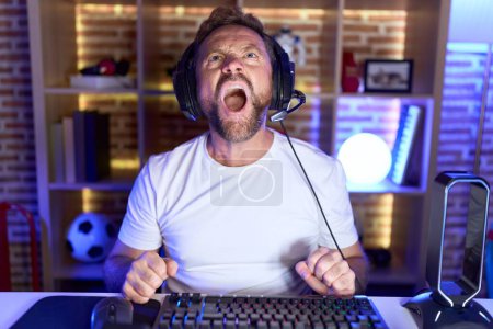 Photo for Middle age man with beard playing video games wearing headphones angry and mad screaming frustrated and furious, shouting with anger. rage and aggressive concept. - Royalty Free Image