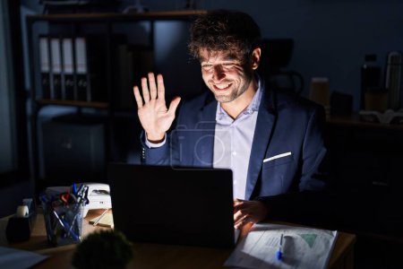 Foto de Joven hispano trabajando en la oficina por la noche mostrando y señalando con los dedos el número cinco mientras sonríe confiado y feliz. - Imagen libre de derechos