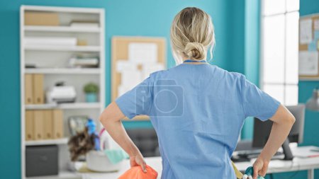 Foto de Joven mujer rubia limpiadora profesional sosteniendo tela y rociador de pie hacia atrás en la oficina - Imagen libre de derechos
