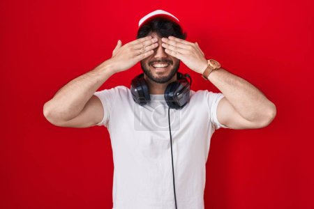Foto de Hombre hispano con barba con sombrero de gamer y auriculares cubriendo los ojos con las manos sonriendo alegre y divertido. concepto ciego. - Imagen libre de derechos