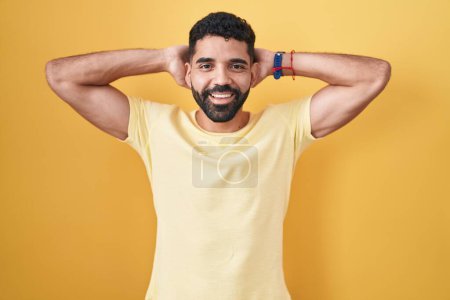Téléchargez les photos : Homme hispanique avec barbe debout sur fond jaune relaxant et étirant, bras et mains derrière la tête et le cou souriant heureux - en image libre de droit