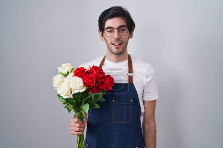 Téléchargez les photos : Jeune homme hispanique tenant bouquet de roses blanches et rouges clignotant en regardant la caméra avec expression sexy, visage joyeux et heureux. - en image libre de droit