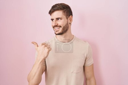 Foto de Hispanic man with beard standing over pink background smiling with happy face looking and pointing to the side with thumb up. - Imagen libre de derechos
