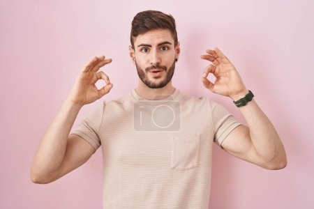 Photo for Hispanic man with beard standing over pink background looking surprised and shocked doing ok approval symbol with fingers. crazy expression - Royalty Free Image
