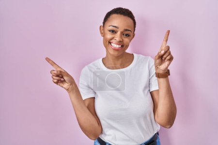 Foto de Hermosa mujer afroamericana de pie sobre fondo rosa sonriendo confiada señalando con los dedos a diferentes direcciones. espacio de copia para publicidad - Imagen libre de derechos