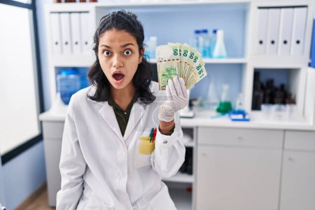 Foto de Mujer hispana con cabello oscuro trabajando en laboratorio científico sosteniendo dinero asustada y sorprendida con la boca abierta para sorpresa, cara de incredulidad - Imagen libre de derechos