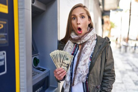 Foto de Mujer rubia joven sosteniendo billetes de dólares de cajero automático asustado y sorprendido con la boca abierta para sorpresa, cara de incredulidad - Imagen libre de derechos