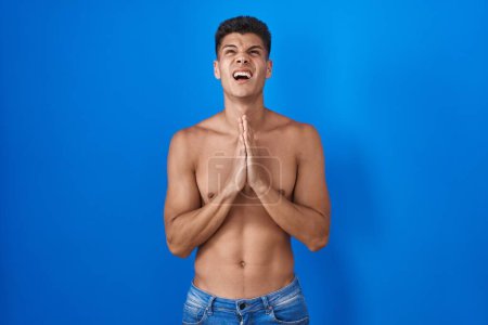 Téléchargez les photos : Jeune homme hispanique debout torse nu sur fond bleu suppliant et priant avec les mains ainsi que l'expression de l'espoir sur le visage très émotionnel et inquiet. mendier. - en image libre de droit