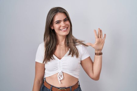 Foto de Joven hermosa mujer con camiseta blanca casual mostrando y apuntando hacia arriba con los dedos número cinco mientras sonríe confiado y feliz. - Imagen libre de derechos