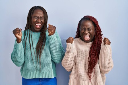Foto de Dos mujeres africanas de pie sobre fondo azul emocionadas por el éxito con los brazos levantados y los ojos cerrados celebrando la victoria sonriendo. concepto ganador. - Imagen libre de derechos