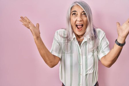 Téléchargez les photos : Femme du Moyen Âge avec les cheveux gris debout sur fond rose célébrant la victoire avec sourire heureux et expression gagnante avec les mains levées - en image libre de droit