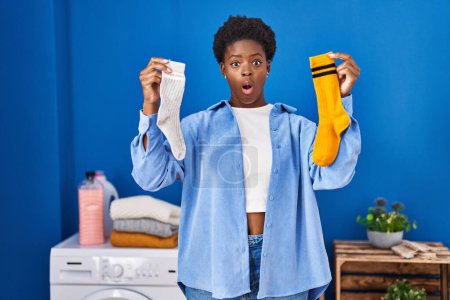 Foto de Mujer afroamericana sosteniendo limpios calcetines sucios Andy miedo y conmocionado con sorpresa y expresión asombrada, miedo y cara emocionada. - Imagen libre de derechos