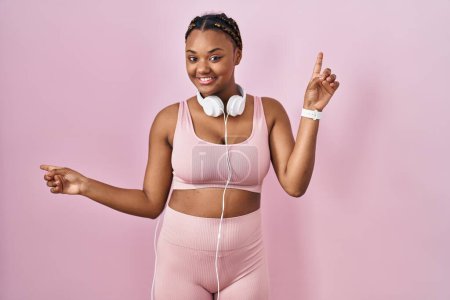 Téléchargez les photos : Femme afro-américaine avec des tresses portant des vêtements de sport et des écouteurs souriant confiant pointant avec les doigts vers différentes directions. espace de copie pour la publicité - en image libre de droit