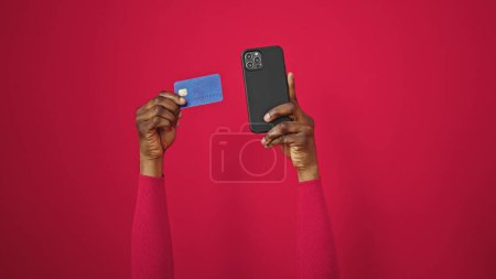 Téléchargez les photos : Femme afro-américaine tenant smartphone et carte de crédit sur fond rouge isolé - en image libre de droit