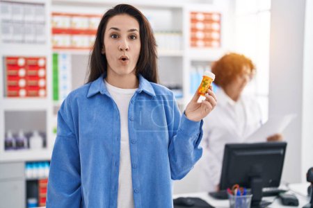 Foto de Joven morena sosteniendo pastillas en la farmacia asustada y sorprendida con la boca abierta para sorpresa, cara de incredulidad - Imagen libre de derechos