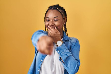 Foto de Mujer afroamericana con trenzas de pie sobre fondo amarillo riéndose de ti, señalando con el dedo a la cámara con la mano sobre la boca, expresión de vergüenza - Imagen libre de derechos