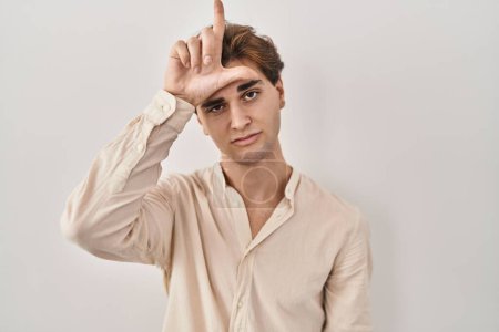 Foto de Young man standing over isolated background making fun of people with fingers on forehead doing loser gesture mocking and insulting. - Imagen libre de derechos