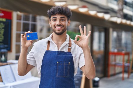 Photo for Arab man with beard wearing waiter apron at restaurant terrace holding credit card doing ok sign with fingers, smiling friendly gesturing excellent symbol - Royalty Free Image