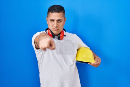 Téléchargez les photos : Jeune homme hispanique tenant un casque pointant du doigt vers la caméra et vers vous, geste confiant et sérieux - en image libre de droit