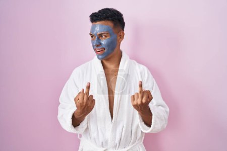 Téléchargez les photos : Young hispanic man wearing beauty face mask and bath robe showing middle finger doing fuck you bad expression, provocation and rude attitude. screaming excited - en image libre de droit