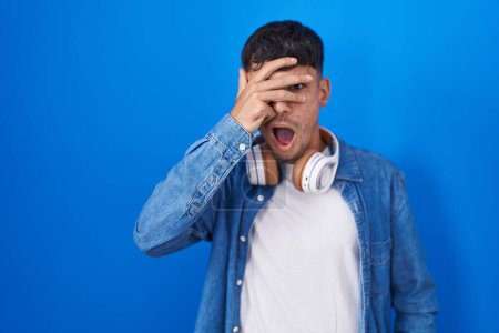 Téléchargez les photos : Young hispanic man standing over blue background peeking in shock covering face and eyes with hand, looking through fingers with embarrassed expression. - en image libre de droit