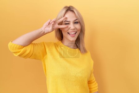 Téléchargez les photos : Jeune femme caucasienne portant un pull jaune faisant symbole de paix avec les doigts sur le visage, souriant joyeuse montrant la victoire - en image libre de droit
