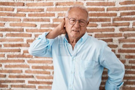 Téléchargez les photos : Homme âgé avec les cheveux gris debout sur le mur de briques souffrant d'une blessure au cou, toucher le cou avec la main, douleur musculaire - en image libre de droit