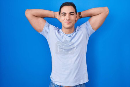Foto de Joven hombre hispano de pie sobre fondo azul relajante y estiramiento, brazos y manos detrás de la cabeza y el cuello sonriendo feliz - Imagen libre de derechos