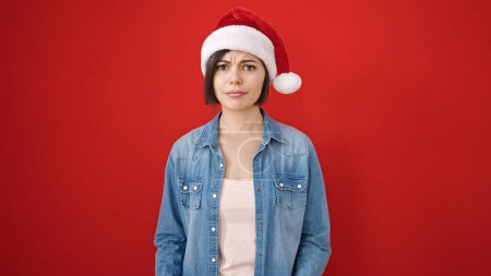 Foto de Mujer caucásica joven de pie con expresión seria usando sombrero de Navidad sobre fondo rojo aislado - Imagen libre de derechos