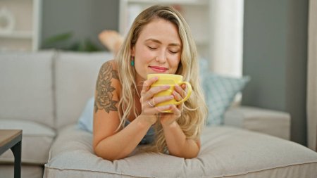 Foto de Mujer rubia joven oliendo taza de café tumbado en el sofá en casa - Imagen libre de derechos