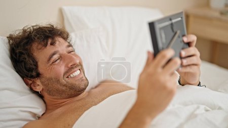 Téléchargez les photos : Jeune homme hispanique allongé sur le lit torse nu regardant photo dans la chambre - en image libre de droit