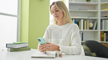Téléchargez les photos : Jeune étudiante blonde utilisant un smartphone à la bibliothèque de l'université - en image libre de droit