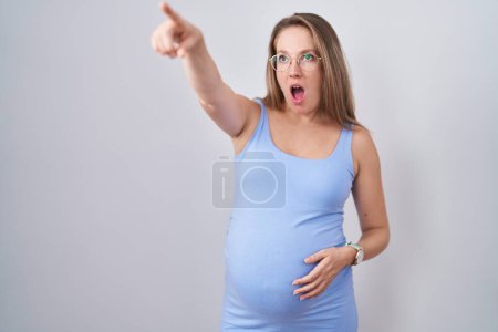 Téléchargez les photos : Jeune femme enceinte debout sur fond blanc pointant du doigt surpris devant, bouche ouverte expression étonnée, quelque chose sur le devant - en image libre de droit