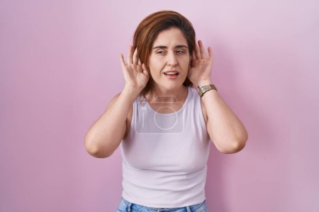 Foto de Mujer morena de pie sobre fondo rosa tratando de escuchar ambas manos en el gesto de la oreja, curioso por chismes. problema auditivo, sordo - Imagen libre de derechos