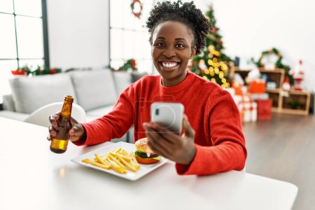 Photo for African american woman eating hamburger using smartphone sitting on table by christmas tree at home - Royalty Free Image
