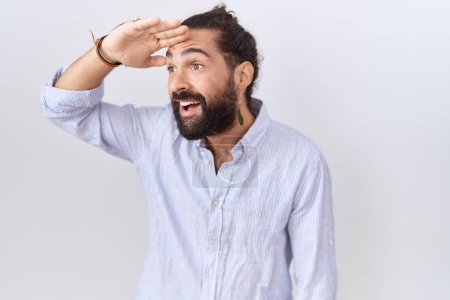 Foto de Hombre hispano con barba vistiendo camisa casual muy feliz y sonriente mirando lejos con la mano sobre la cabeza. concepto de búsqueda. - Imagen libre de derechos