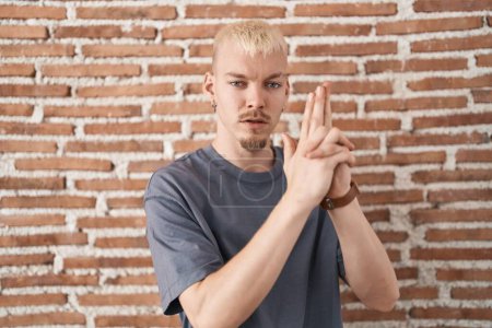 Photo for Young caucasian man standing over bricks wall holding symbolic gun with hand gesture, playing killing shooting weapons, angry face - Royalty Free Image